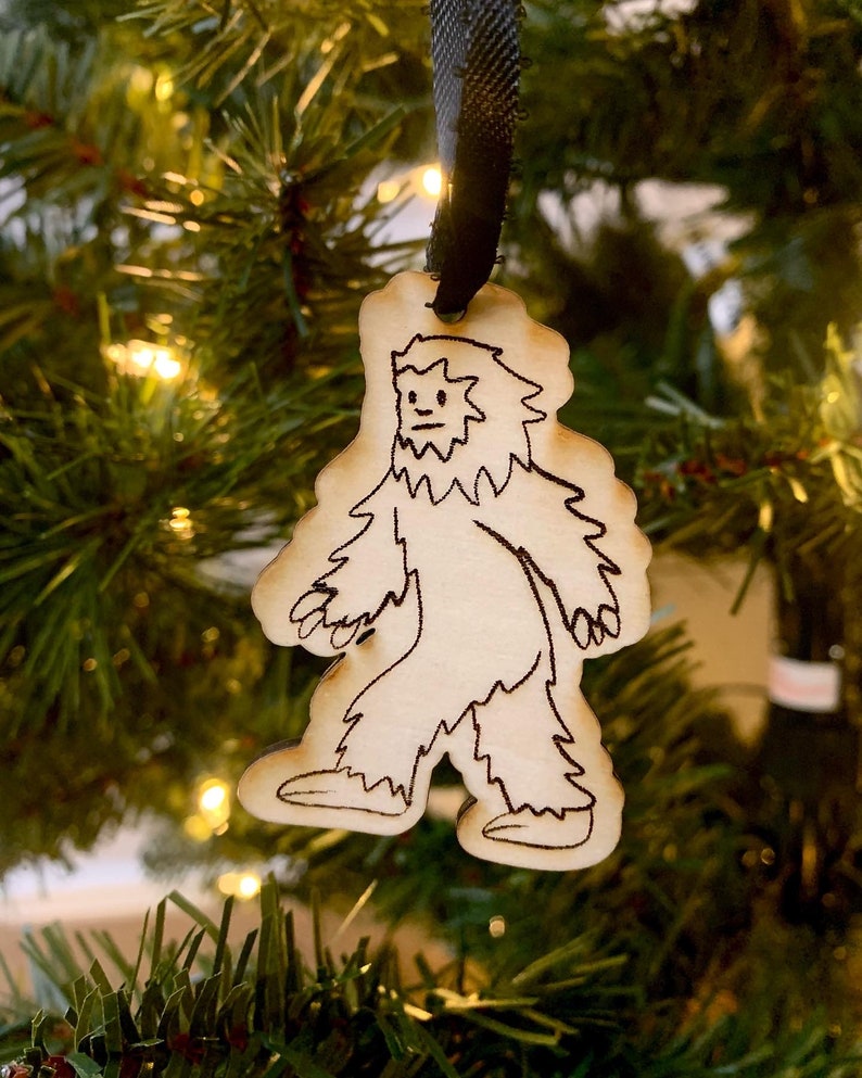 May include: A wooden Christmas ornament shaped like a Bigfoot, with a black ribbon attached. The ornament is hand-drawn in black ink and features a simple design of a Bigfoot walking.