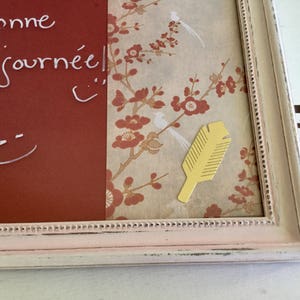 Peut inclure: Un tableau noir encadré blanc avec une bordure rose et un motif floral sur le côté droit. Le tableau noir porte les mots "Bonne journee!" écrits à la craie blanche. Une plume en papier jaune est fixée au motif floral.