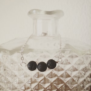 May include: A silver chain necklace featuring three dark, textured, round beads. The necklace is displayed against a clear, diamond-patterned glass bottle, creating a minimalist aesthetic. The beads appear to be lava rock.