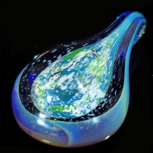 May include: A teardrop-shaped glass pendant featuring a swirling pattern of deep blue, bright green, and white colors. The surface has a smooth, polished finish with translucent layers that create depth, while the outer edges transition into a lighter, iridescent blue hue against a dark background.