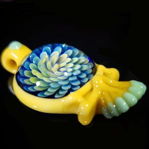 May include: A glass pendant featuring a central circular inlay with a blue and light green spiral floral pattern. The surrounding frame is made of opaque yellow glass with a smooth finish, including a bail at the top and a textured tail section.