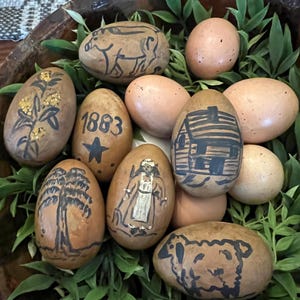 May include: A collection of brown eggs decorated with various hand-painted designs, including a pig, a house, a tree, a woman, a sheep, and the year 1883. The eggs are nestled in a bowl with green leaves.