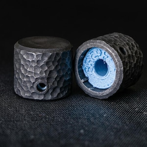 May include: Two cylindrical knobs featuring a dark gray, hammered metal exterior texture. Each knob has a small circular hole on the side. One knob is positioned upright, while the other is tilted to reveal a bright blue, textured plastic interior.