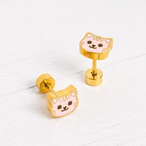 May include: A pair of gold-tone stud earrings featuring a flat, cat-shaped face with light pink enamel detailing, black eyes, and a small black nose. The earrings have a cylindrical gold-tone post backing and a smooth, polished metallic finish.