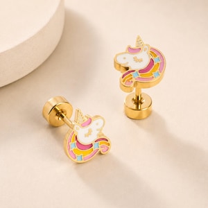 May include: A pair of gold-tone stud earrings featuring a unicorn head design. The unicorn is filled with white, pink, and yellow enamel, accented with small blue star shapes. Each earring has a round gold-tone backing post and a secure screw-back.