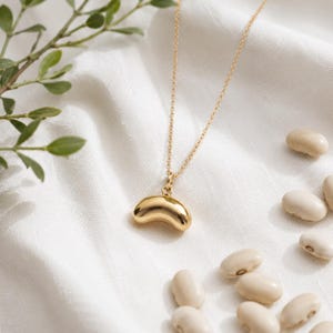 Op de afbeelding: Een goudkleurige ketting met een gladde, gebogen, boonvormige hanger aan een fijne ketting. De hanger heeft een gepolijst, reflecterend oppervlak dat het licht opvangt, wat zorgt voor een eenvoudige en strakke uitstraling van dit sieraad.