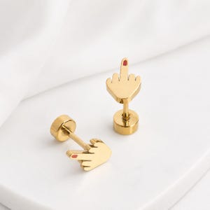 May include: A pair of gold-toned stud earrings shaped like a hand making a middle finger gesture. Each earring features a small red enamel detail on the tip of the middle finger and a circular backing post, crafted with a polished metallic finish.