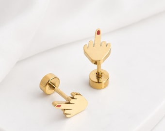 Pode incluir: Um par de brincos de pino dourados em forma de mão fazendo um gesto de dedo médio. Cada brinco apresenta um pequeno detalhe em esmalte vermelho na ponta do dedo médio e um fecho circular, com um acabamento metálico polido.