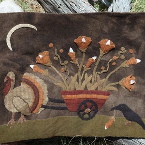 May include: A brown felt wall hanging depicting a turkey, a crow, and a cart of flowers. The turkey has a red wattle and a fan of orange, brown, and tan feathers. A crescent moon is in the upper left corner.