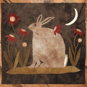 May include: A brown rabbit sits in a field of red flowers with a crescent moon in the background. The rabbit is made of fabric and is stitched with brown thread. The flowers are also made of fabric and are stitched with red thread. The background is a dark brown fabric.