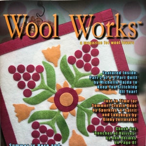 May include: The cover of "Wool Works" magazine features a white square with a floral design in orange, green, and burgundy. The magazine's title is in orange, and the cover includes text about summer projects and featured articles.