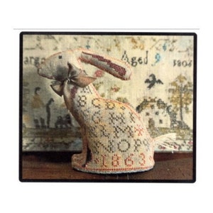 May include: A vintage needlepoint rabbit with a red, white, and blue bow. The rabbit is covered in embroidered letters and numbers. The rabbit is sitting on a table in front of a needlepoint sampler with the text "Aged 9 1804".