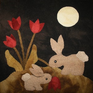 May include: Two brown rabbits with black eyes sit in front of a patch of red tulips under a full moon. The background is a dark brown fabric.