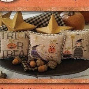 May include: A collection of six Halloween-themed pincushions. The pincushions are made of fabric and feature various Halloween designs, including ghosts, goblins, trick or treat, and a witch. The pincushions are displayed on a wooden tray with a black and white checkered cloth.