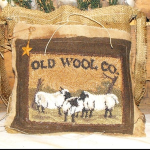 Può includere: Un sacco di iuta rustico con una toppa rettangolare con la scritta "OLD WOOL CO." e un'immagine di pecore. La borsa è riempita di verde e soffici accenti bianchi. Un piccolo ciondolo a forma di stella dorata pende dalla borsa.