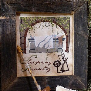 May include: A framed cross-stitch embroidery depicting a scene from the fairy tale Sleeping Beauty. The embroidery shows a sleeping princess in a stone alcove with a spinning wheel and the text "Sleeping Beauty".