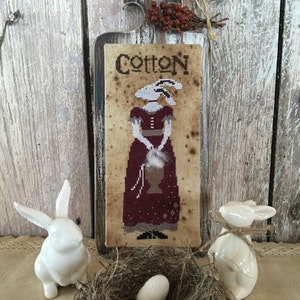 May include: A rustic wooden sign with the word "Cotton" and a cross-stitch design of a woman in a burgundy dress. Two white ceramic bunny figurines flank a small nest with an egg. The sign is adorned with a sprig of red berries.