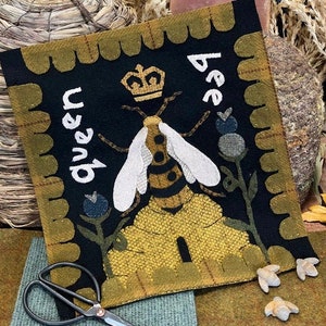 May include: A black felt wall hanging with a bee and honeycomb design. The bee is wearing a crown and the text "bee queen" is written on the hanging.
