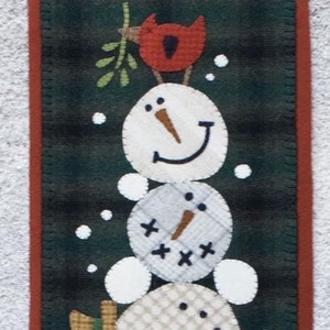 Può includere: Un arazzo da parete in feltro con una pila di cinque pupazzi di neve su uno sfondo verde con fiocchi di neve bianchi. I pupazzi di neve sono fatti di feltro bianco con occhi e bocca neri, nasi arancioni e bastoncini marroni per le braccia. Il pupazzo di neve in cima ha un cardinale rosso appollaiato sulla testa.