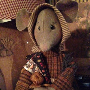 May include: A gray and brown striped fabric mouse doll holding a bundle of yarn and fabric scraps. The doll is wearing a brown and white striped fabric bonnet. The doll is sitting on a brown and white checkered fabric surface.