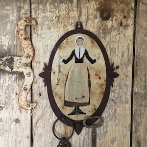 May include: A framed cross-stitch of a woman holding keys, with two antique keys hanging below the frame on a wooden background.