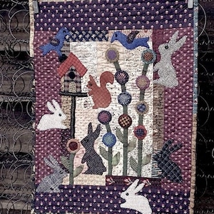 May include: A quilted wall hanging with a purple background and a whimsical design featuring bunnies, birds, squirrels, and flowers. The text "Hoppity Lane... by: Marian Edwards" is stitched into the bottom right corner.