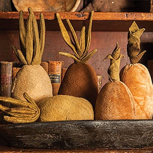 May include: A wooden bowl filled with five handmade felt pineapples in shades of brown and tan. The pineapples are arranged in a cluster, with their tops facing up. The bowl is sitting on a wooden shelf with books and other items in the background.