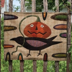 May include: A brown burlap wall hanging with a black crow carrying a smiling orange pumpkin. The background is decorated with colorful, abstract shapes.