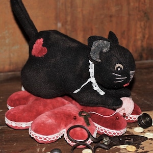 May include: A black felt cat pincushion with a red heart on its side, sitting on a red velvet heart-shaped pincushion with white lace trim. The cat is holding a pink ball of yarn and there are sewing supplies scattered around it, including scissors, a thimble, and buttons.