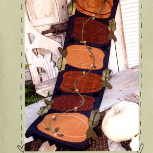 May include: A wool-appliqued table runner featuring a design of five pumpkins in shades of orange, brown, and black, with a vine of green leaves and small white flowers. The runner is 12 inches by 44 inches and is titled "#325 Pumpkin Harvest".