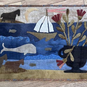 May include: A handmade textile art piece depicting a seascape with a sailboat, whale, and horses. A black vase holds flowers with red and brown accents. The artwork has a brown border and is set against a neutral background.