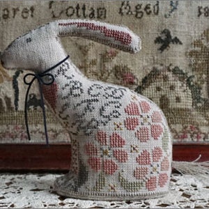 May include: A white stuffed rabbit with a long, floppy ear, sitting on a floral patterned fabric. The rabbit has a black ribbon around its neck and is embroidered with pink and green flowers.