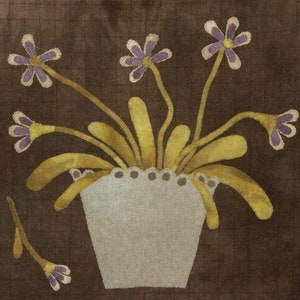 May include: A brown fabric background with a white ceramic pot with a scalloped rim. The pot is filled with yellow stems and purple flowers.