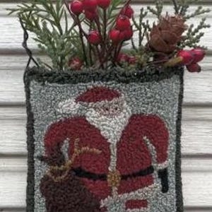 May include: A hand-stitched wool rug depicting Santa Claus carrying a sack of gifts. The rug is framed in a dark green border and is hanging from a wire hanger. The rug is surrounded by a sprig of greenery with red berries.