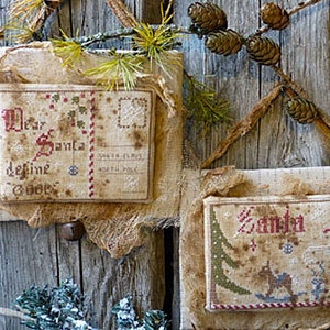 May include: Two vintage-style Christmas ornaments with cross-stitch designs. The first ornament is a letter to Santa with the text "Dear Santa define good". The second ornament says "Santa" with a cross-stitch image of Santa Claus and a Christmas tree.