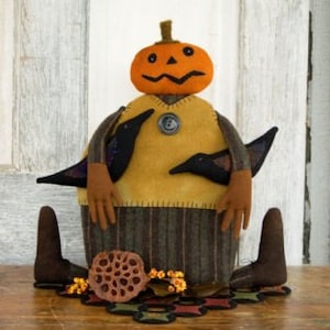 May include: A handmade felt pumpkin doll with a yellow sweater, black birds, and brown boots. The doll is sitting on a brown surface with a decorative leaf and berries.