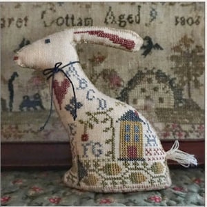 May include: A white fabric bunny with embroidered details, including a red heart, a house, and a floral pattern. The bunny is sitting on a textured surface.