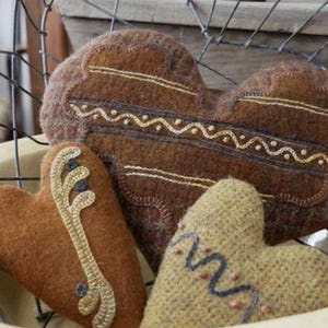 May include: Three heart-shaped decorative pillows in a wire basket. The pillows are brown, tan, and beige, with embroidered details in gold, blue, and cream. The pillows have a rustic, handmade appearance.