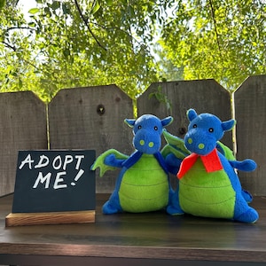 May include: Two plush blue and green dragon toys with outstretched wings and a small red scarf. A small chalkboard sign with the words "ADOPT ME!" stands in front of the dragons on a wooden surface.