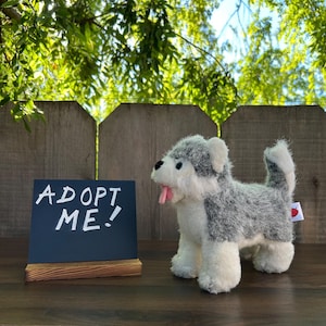 May include: A plush grey and white husky dog toy with a pink heart on its back. The toy is sitting on a wooden table with a black chalkboard sign that says "Adopt Me!"