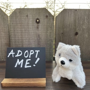 May include: A gray and white plush wolf toy sits next to a small chalkboard sign that reads "ADOPT ME!" in white lettering. The sign is on a wooden base. The background features a weathered wooden fence.