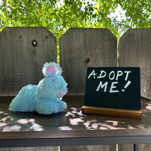May include: A light blue plush bunny toy with a pink nose and ears, lying on a wooden surface. A small chalkboard sign next to the bunny reads "ADOPT ME!" in white chalk. The background includes a wooden fence and green foliage.