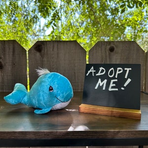 May include: A blue plush whale toy with a white belly and a black eye. The whale is sitting on a wooden table with a black chalkboard sign that says "ADOPT ME!" in white chalk.