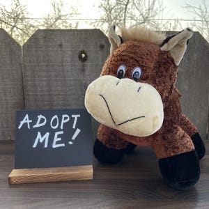 May include: A plush brown horse toy with a cream-colored muzzle and black hooves. The toy is positioned next to a small chalkboard sign that reads "ADOPT ME!" in white chalk. The background includes a wooden fence and a blurred outdoor scene.
