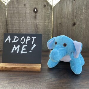 May include: A light blue plush elephant toy with large ears and a trunk. Next to the elephant is a small chalkboard sign that reads "ADOPT ME!" in white lettering. The sign is on a wooden stand.