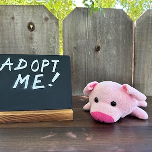 May include: A pink plush pig toy next to a small chalkboard sign that reads "ADOPT ME!" in white letters. The sign is on a wooden base. The background features a wooden fence.