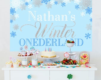 May include: A light blue backdrop featuring silver and blue snowflakes with the text Nathan's Winter ONEDERLAND in white and silver glitter script. The display is set up on a white table with a cake, cake pops, fruit, and colorful candies.