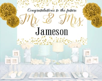 May include: A white rectangular backdrop featuring gold confetti patterns and the black and gold text Congratulations to the future Mr & Mrs Jameson, flanked by four gold paper pom-poms, displayed above a table with cakes and treats.