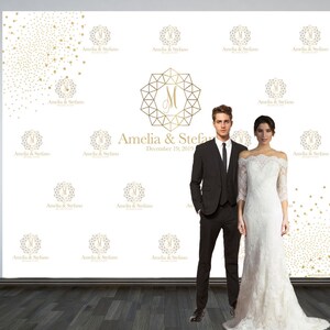 May include: A white backdrop with gold confetti and the text "Amelia & Stefano" repeated multiple times. A large gold geometric monogram with the letter "M" is in the center. The text "December 19, 2019" is below the monogram. A couple is standing in front of the backdrop. The woman is wearing a white off-the-shoulder wedding dress. The man is wearing a black suit and tie.