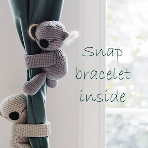 Può includere: Due fermagli per tende a forma di koala in crochet grigio. I koala sono appesi a una tenda verde. Il testo "Snap bracelet inside" è scritto in verde su sfondo bianco.